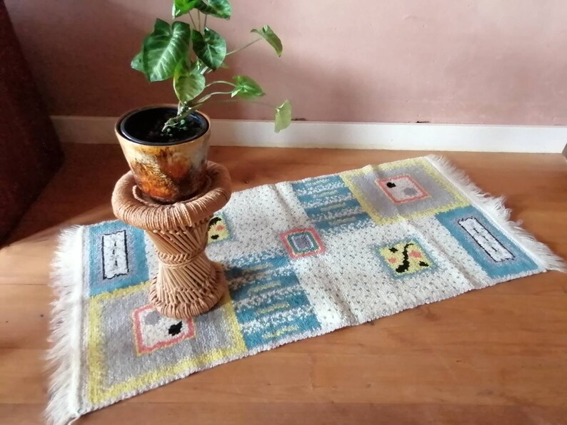 May include: A white rug with a geometric pattern in shades of blue, yellow, and pink. The rug has a fringe border and is placed on a wooden floor. A potted plant with green leaves sits on a wicker stool.