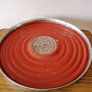 May include: A round, red metal tray with a silver rim. The tray has concentric circular ridges and a central, slightly raised area. The surface shows signs of wear, with some paint loss. The tray sits on a wooden surface.