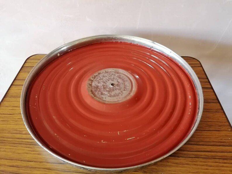 May include: A round, red metal tray with a silver rim. The tray has concentric circular ridges and a central, slightly raised area. The surface shows signs of wear, with some paint loss. The tray sits on a wooden surface.