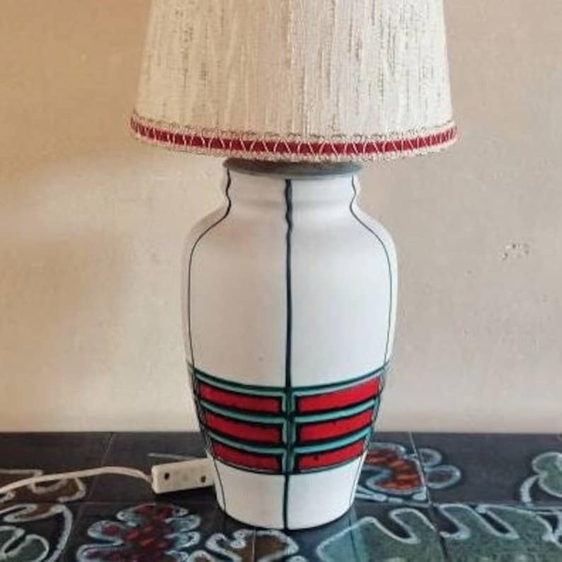 May include: A white ceramic table lamp with a cream-colored lampshade. The lamp base features black vertical lines and red and teal horizontal bands. The lampshade is trimmed with red. The lamp is plugged into an outlet.