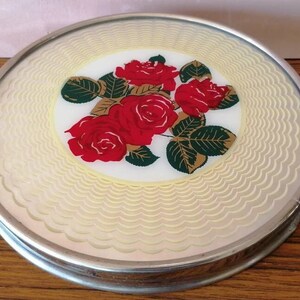 May include: A round, decorative trivet with a silver-colored metal rim. The center features a floral design of red roses, green leaves, and gold accents on a white background. The outer edge has a cream-colored, wavy pattern.