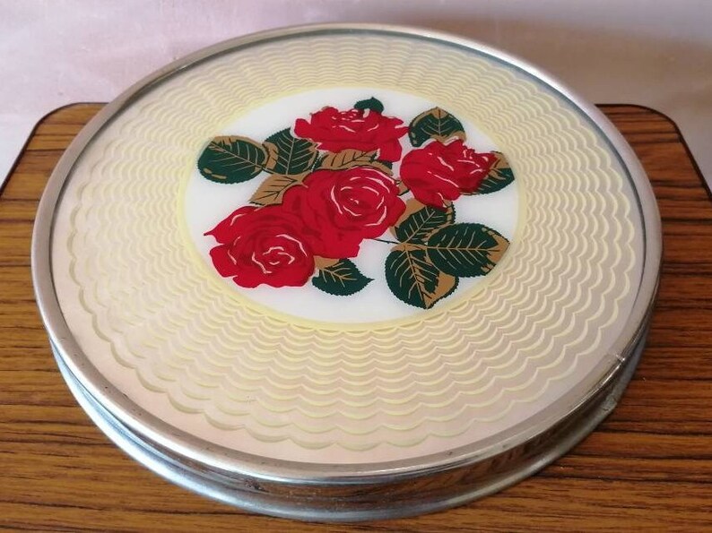 May include: A round, decorative trivet with a silver-colored metal rim. The center features a floral design of red roses, green leaves, and gold accents on a white background. The outer edge has a cream-colored, wavy pattern.