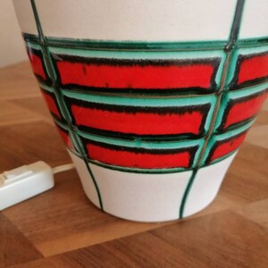 May include: A white ceramic lamp with a geometric design. The lamp features a red and green pattern with black outlines. The lamp has a white cord and a switch. The lamp is on a wooden surface.