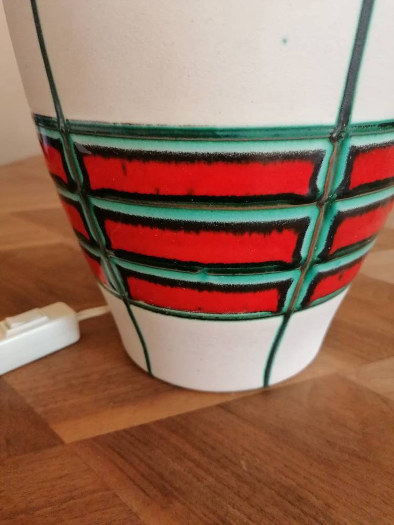 May include: A white ceramic lamp with a geometric design. The lamp features a red and green pattern with black outlines. The lamp has a white cord and a switch. The lamp is on a wooden surface.