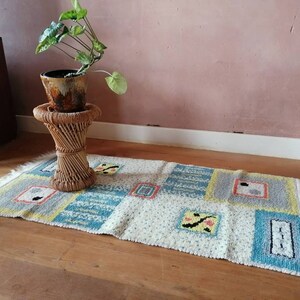 May include: A white and blue patterned rug with fringe on the edges. The rug is on a wooden floor and there is a potted plant on a wicker stool in the background.