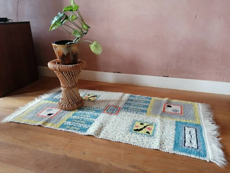May include: A white and blue patterned rug with fringe on the edges. The rug is on a wooden floor and there is a potted plant on a wicker stool in the background.