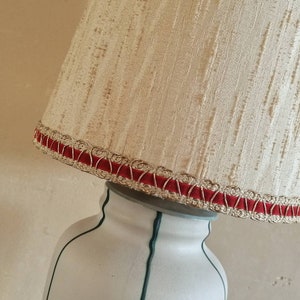 May include: Cream-colored lampshade with a textured surface, trimmed with red velvet and gold-colored trim. The lampshade sits atop a white lamp base with green accents. The lamp is angled.