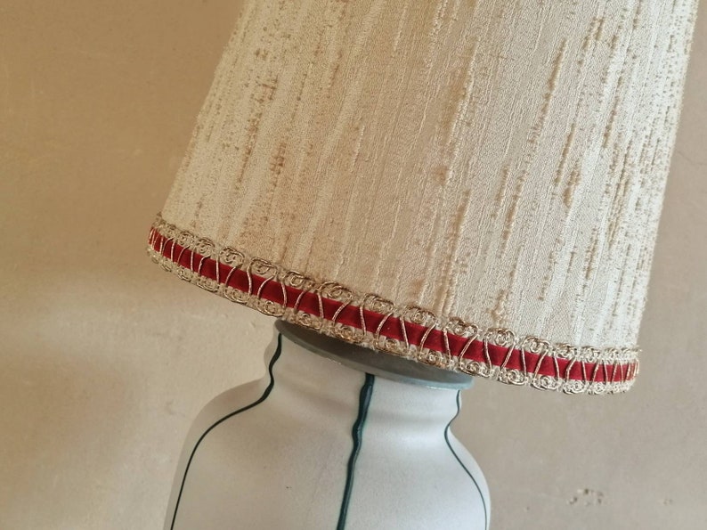 May include: Cream-colored lampshade with a textured surface, trimmed with red velvet and gold-colored trim. The lampshade sits atop a white lamp base with green accents. The lamp is angled.