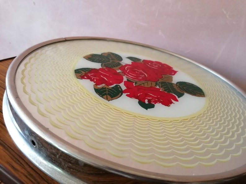 May include: Oval-shaped, vintage vanity tray with a silver-toned metal frame. The tray features a central design of red roses with green leaves on a white background. The outer edge has a scalloped, cream-colored pattern.