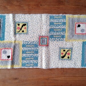 May include: A white rug with a geometric pattern in shades of blue, yellow, gray, and pink. The rug has a fringe border.