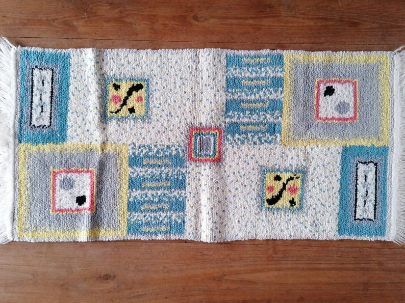 May include: A white rug with a geometric pattern in shades of blue, yellow, gray, and pink. The rug has a fringe border.