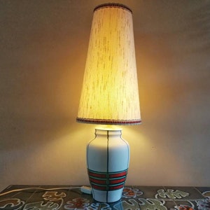 May include: A vintage table lamp with a white ceramic base featuring green and red stripes and a beige lampshade. The lampshade is tall and conical, with a decorative trim at the top and bottom. The lamp is turned on, casting a warm glow.