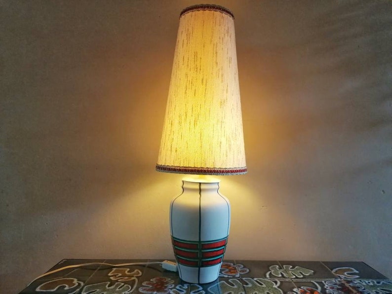 May include: A vintage table lamp with a white ceramic base featuring green and red stripes and a beige lampshade. The lampshade is tall and conical, with a decorative trim at the top and bottom. The lamp is turned on, casting a warm glow.