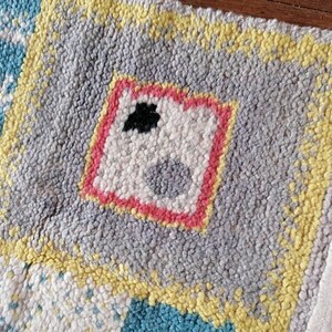 May include: A colorful rug with a geometric design. The rug is made of a soft, fuzzy material and has a fringe border. The rug features a central square with a pink border and a white interior with a black and gray design.