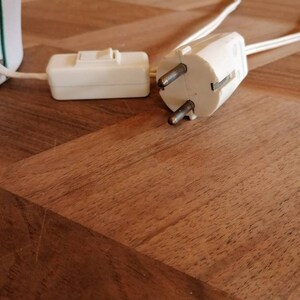 May include: A white electrical plug with two prongs and a ground pin, connected to a white cord. A white inline switch is also visible. The items are on a wooden surface. Electrical components.