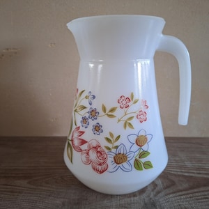 Könnte beinhalten: Weißer Glaskrug mit floralem Muster. Der Krug hat einen Griff und eine weite Öffnung. Das florale Design zeigt rote, blaue und weiße Blumen mit grünen Blättern. Er wird wahrscheinlich zum Servieren von Getränken verwendet.