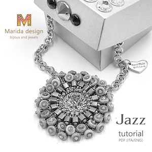 Könnte beinhalten: Eine silberne Halskette mit einem runden Anhänger aus grauen und weißen Perlen. Der Anhänger hat ein Sonnenstrahlen-Design mit einem klaren Kristall in der Mitte. Die Halskette befindet sich in einer Schachtel mit dem Text "Marida design bijoux and jewels" und "Hand Made With Love". Der Text "Jazz tutorial PDF (ITA/ENG)" ist ebenfalls sichtbar.