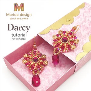 Può includere: Un paio di orecchini con perline rosa e dorate con un pendente a goccia di cristallo. Gli orecchini sono in una piccola scatola rosa con bordatura dorata. La scatola ha un motivo floreale all'interno.