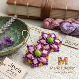 Könnte beinhalten: Ein Paar perlenbesetzte Blumenohrringe mit einer zentralen lindgrünen Perle, umgeben von hellrosa und dunkelvioletten Blütenblättern, akzentuiert mit kleinen gelben Kristallen. Die Ohrringe tragen die Aufschrift Myrta Marida design.