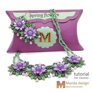 Può includere: Una collana e orecchini di perline viola e verdi con un design a forma di fiore. I gioielli sono esposti in una scatola a cuscino rosa con il testo "Spring flowers" e "tutorial PDF (ITA/ENG) Marida design bijoux and jewels".