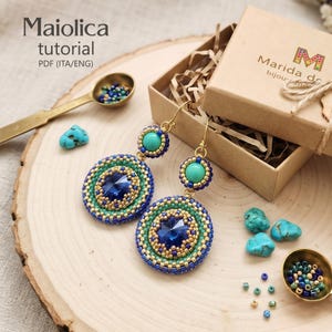 May include: Beaded drop earrings featuring a central dark blue faceted crystal surrounded by concentric rings of turquoise, gold, and blue seed beads. The earrings include a smaller turquoise bead at the top and gold-tone hooks. Text reads Maiolica tutorial PDF (ITA/ENG) and Marida de bijoux.