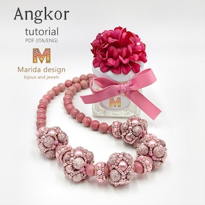 Può includere: Una collana di perline con sfere color rosa polvere e complessi grappoli di perline testurizzati a forma di fiore nei toni del rosa e del malva. Dietro la collana si trova un piccolo barattolo di vetro sormontato da un fiore in tessuto rosa.