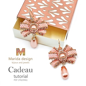 May include: A pair of earrings featuring a bow design crafted from small light pink beads with metallic gold accents. Each earring includes a central round bead and a dangling teardrop-shaped pearl-like bead. Text reads: M Marida design bijoux and jewels Cadeau tutorial PDF (ITA/ENG).