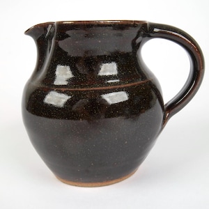 May include: A dark brown ceramic pitcher with a glossy, speckled glaze finish. The vessel features a rounded body, a wide pouring spout, and a curved handle on the right side. The base of the pitcher shows a natural, unglazed clay rim.