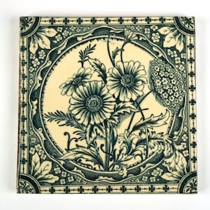 May include: A square ceramic tile featuring a dark teal floral pattern on a cream background. The design includes a central circular medallion filled with detailed daisies and foliage, framed by a decorative border of small flowers and leaves.