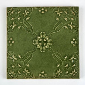 May include: A square ceramic tile in a deep olive green colour featuring a raised relief pattern. The design consists of a central four-petaled floral motif with symmetrical, flowing vine-like stems extending toward each corner of the tile.