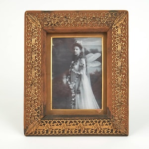 Pode incluir: Uma moldura retangular com borda de madeira marrom escura e uma sobreposição ornamental de filigrana em tom dourado metálico com padrões florais e de folhas. A moldura contém uma fotografia em preto e branco de uma figura com asas.