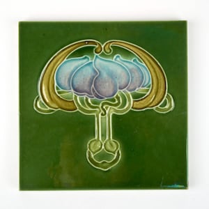 May include: Square green ceramic tile with a floral design. The design features a stylized blue and purple flower with gold and green accents. The tile has a glossy finish and is likely for decorative use.