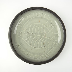 May include: Round ceramic plate with a light green center and a dark brown rim. The plate features an embossed design of two stylized fish facing each other. The plate is a decorative item.