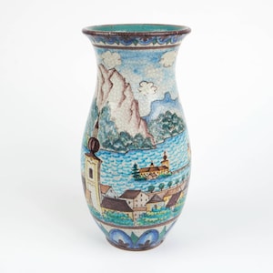 Könnte beinhalten: Eine Keramikvase mit ausgestelltem Rand und einer rissigen Glasuroberfläche. Die Außenseite zeigt eine handgemalte Landschaftsszene mit einer Stadt am See, einer Kirche, Bergen und blauem Wasser, akzentuiert durch grüne Bäume und braune Dächer.