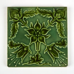 Puede incluir: Un azulejo de cerámica cuadrado con un esmalte verde salvia profundo que presenta un patrón de relieve simétrico de motivos florales estilizados, hojas y zarcillos delicados que se extienden hacia las esquinas con un acabado brillante.