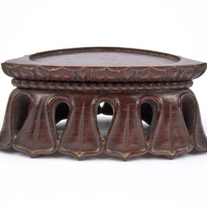 May include: A dark brown, carved wooden display stand with a rectangular top and a decorative, scalloped edge. The stand features a detailed, openwork design with curved supports and a raised, circular base. The wood has a rich, reddish-brown hue.