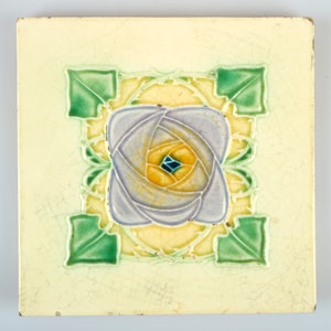 May include: A square ceramic tile with a raised floral design. The flower is a stylized rose with a purple center and yellow petals. The background is a pale yellow with green leaves around the flower.