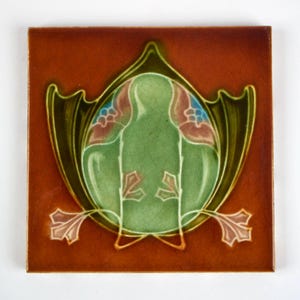 May include: A square ceramic tile with a glossy finish featuring a deep reddish-brown background. Centered is a stylized green oval motif surrounded by dark green leaf-like shapes, accented with small pink and blue floral details at the top.