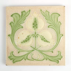 Puede incluir: Un azulejo de cerámica blanco con un diseño floral verde. El azulejo presenta tres flores estilizadas con hojas y tallos verdes. El azulejo tiene un esmalte craquelado.