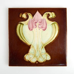 May include: Square brown ceramic tile with a floral design in pink, green, and gold. The Art Nouveau style tile features a central pink flower shape with green and gold accents, set against a dark brown background.