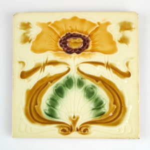 May include: Square ceramic tile featuring a stylized floral design. The central flower is a vibrant orange with a dark purple center. Green and brown foliage accents the design against a cream-colored background. The tile has a glossy finish.