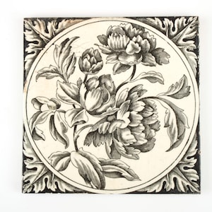 Puede incluir: Azulejo cerámico cuadrado con diseño floral. La imagen central presenta una ilustración detallada en blanco y negro de peonías y hojas dentro de un marco circular. El borde es negro con un patrón de hojas repetido.
