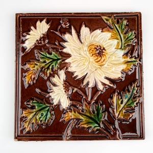 Könnte beinhalten: Quadratische, braune Keramikfliese mit erhabenem Blumendesign. Die Fliese zeigt cremefarbene Blüten mit braunen Zentren und grünen und gelben Blättern. Die Fliese hat einen erhabenen Rand.