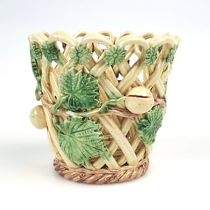 Op de afbeelding: Een keramische plantenpot in de vorm van een gevlochten mand met groene bladeren en witte bessen. De pot is versierd met een bruine touwachtige rand rond de bovenkant.