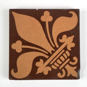 May include: A square ceramic tile featuring a dark brown background with a contrasting light brown fleur-de-lis pattern centred on the surface. The design includes stylised leaf shapes and a base element with horizontal lines, creating a flat graphic.