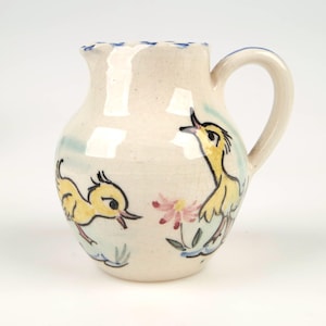May include: A white ceramic jug with a blue rim and a handle. The jug is decorated with hand-painted yellow chicks and pink flowers.