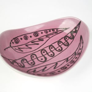 May include: A pink ceramic serving dish with a white rim. The dish features three black feather designs with intricate patterns. The dish has a smooth, glossy finish and is set against a white background.