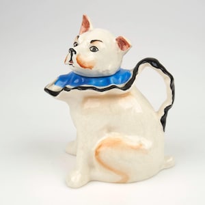 Op de afbeelding: Een keramische theepot in de vorm van een hond. De theepot is wit met een blauwe kraag en een zwarte handgreep. De hond heeft bruine oren en een bruine neus. De theepot staat op een witte ondergrond.