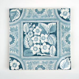 May include: Square ceramic tile with a floral design in shades of blue and white. The central square features white flowers and green leaves, framed by a border with floral and leaf motifs. The corners have stylized flower designs.