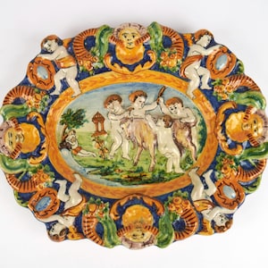 Könnte beinhalten: Eine ovale Keramikplatte mit einer handbemalten Szene in der Mitte, die Putten zeigt, die mit einer Ziege in einer Landschaft interagieren. Der Rand ist mit reliefartigen Putten, sonnenartigen Gesichtern und Blumenmotiven in Blau, Orange und Grün verziert.